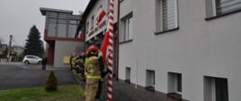 Na zdjęciu widzimy na pierwszym planie strażaków w pełnym uzbrojeniu, którzy dokonują podniesienia Flagi państwowej na maszt. Na drugim planie widać pozostałych strażaków stojących w szeregu oraz komendanta. W tle widać budynek komendy. 