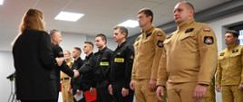 W sali uroczystości komendant wręcza dokumenty. Przed nimi stoi szereg strażaków w czarnych i piaskowych mundurach, ustawionych w formalnym szyku. Kilka osób trzyma czerwone teczki, a scena wygląda na moment wręczania wyróżnień.