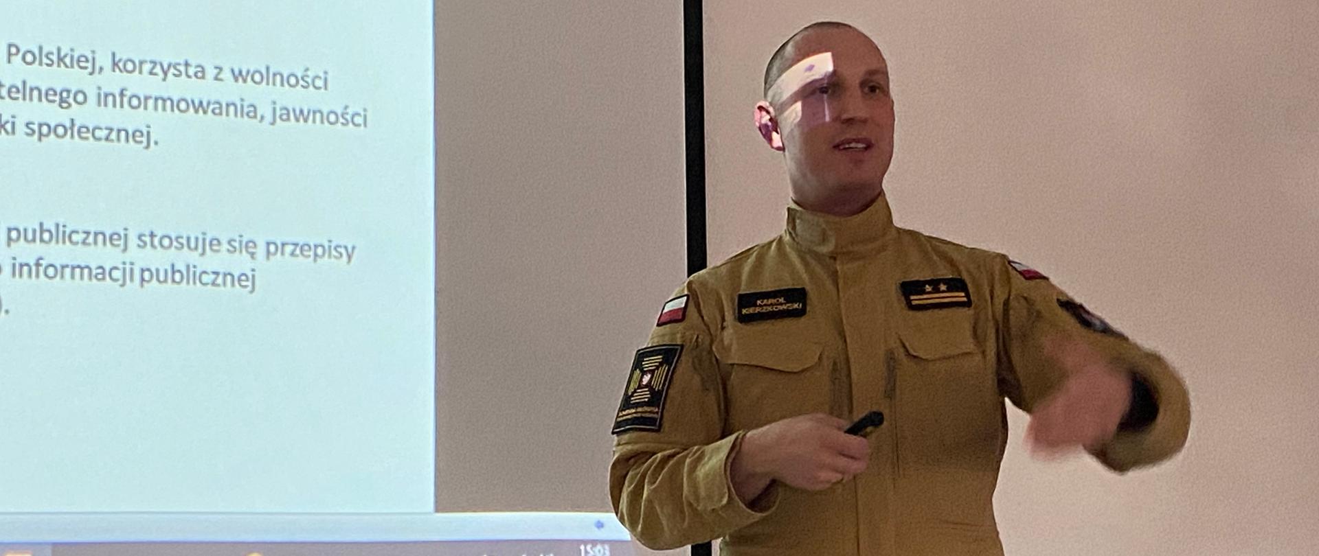 Strażaka w umundurowaniu służbowym przemawia do strażaków na szkoleniu. W tle ekran z wyświetlaną treścią prezentacji.