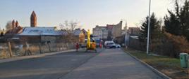 Ulica Wodna w Jutrosinie. Na jezdni stoi smigłowiec Lotniczego Pogotowia Ratunkowego. Z jego lewej strony znajduje się członek załogi. Z prawej strony cześciowo na parkingu zaparkowana jest karetka pogotowia, kóra ma włączone światła błyskowe. Za nia widoczny jest wóz strażacki. Pomiędzy pojazdami znajdują się strażacy i ratownik medyczny. W tle kamienice, z lewej strony natomiast widać wieże kościoła.