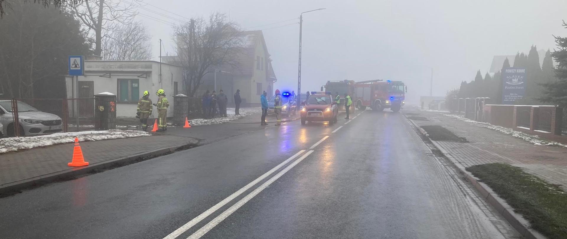 Na drodze stoją pojazdy pożarnicze podczas działań. Panuje mgła.