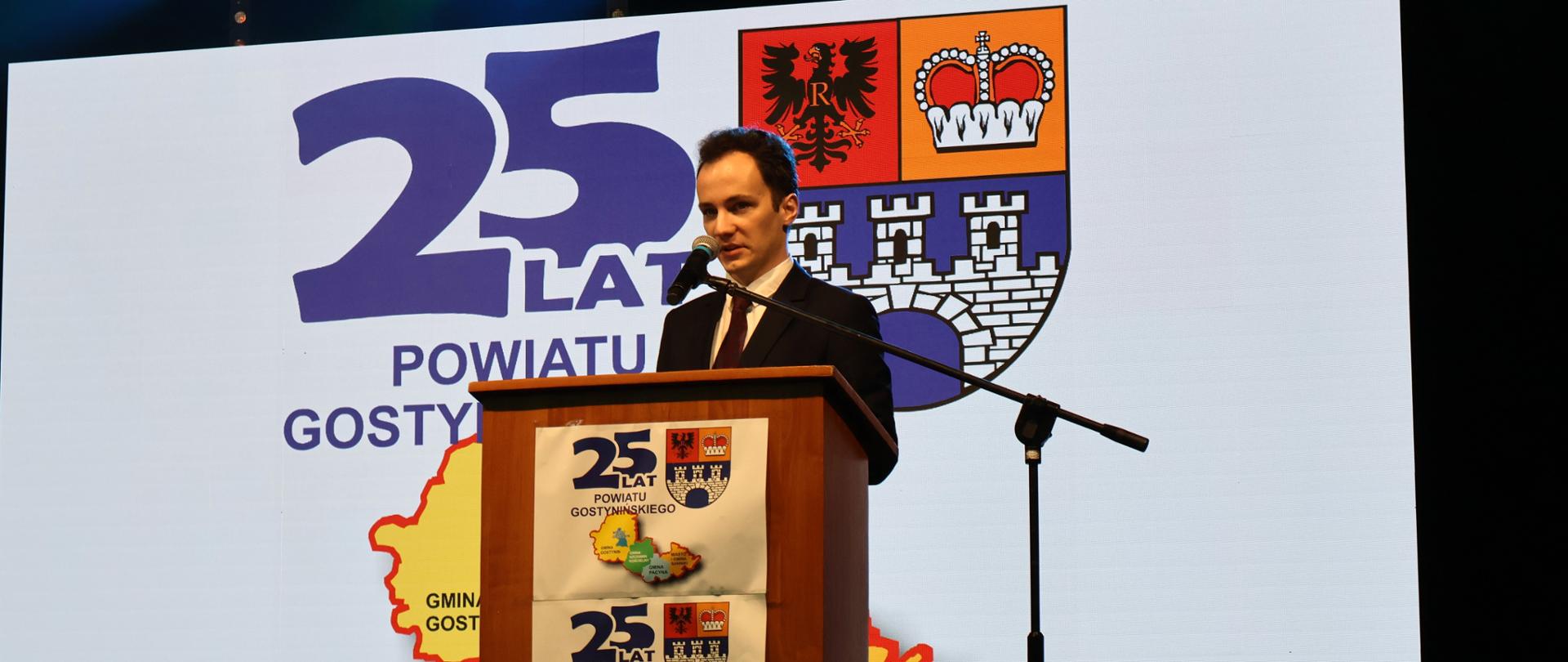 Na zdjęciu mężczyzna stojący przy mównicy. W tle napis 25 - lecie Powiatu Gostynińskiego 