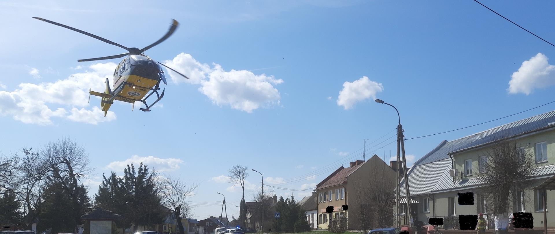 Zdjęcie obrazuje kilka samochodów osobowych ciągnik rolniczy lądujący śmigłowiec LPR, radiowóz, strażaka, budynki i drzewa w tle