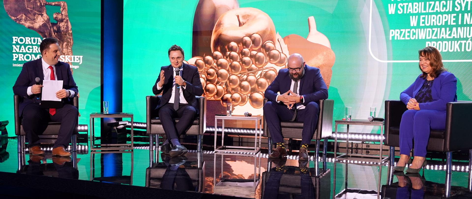 podsekretarz stanu Krzysztof Ciecióra siedzący wśród prelegentów z mikrofonem w dłoni