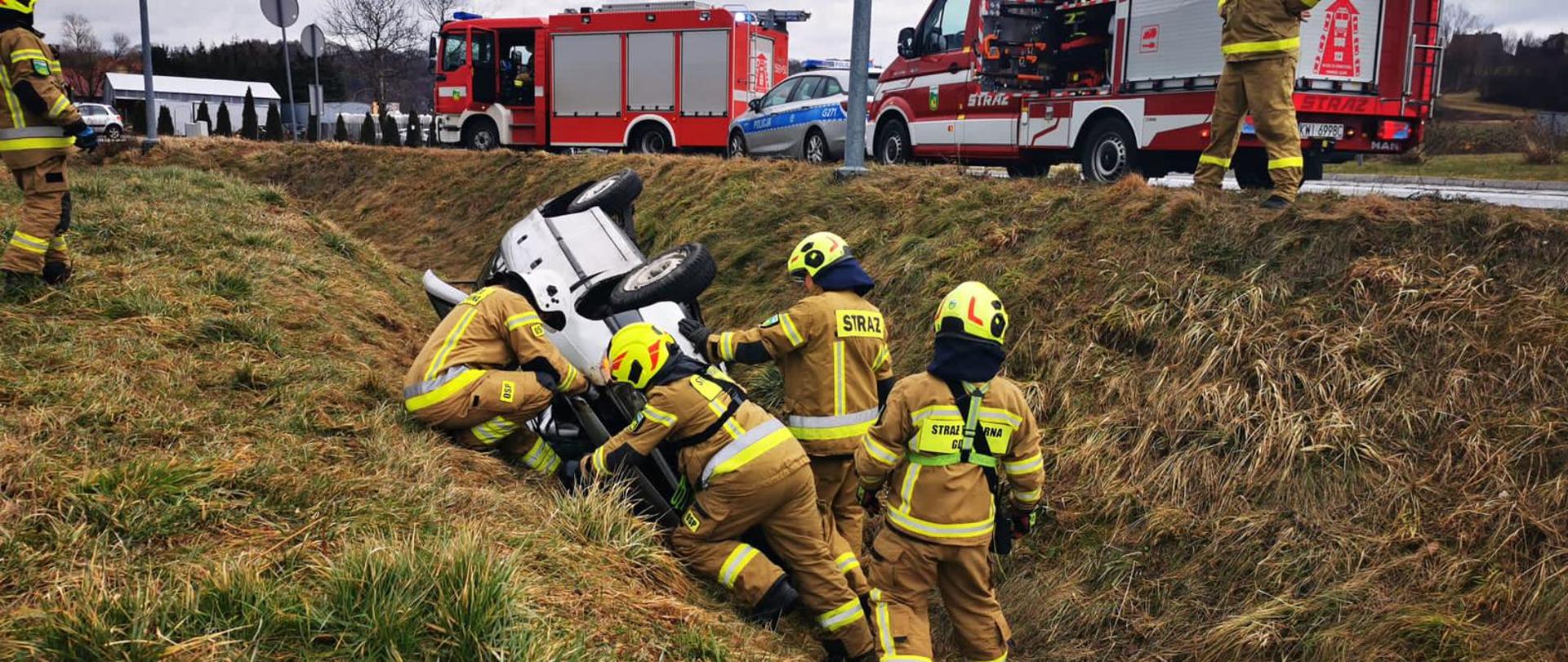 Na zdjęciu samochód osobowy na dachu poza drogą. Przy samochodzie strażacy w ubraniach specjalnych prowadzą działania. W tle samochody pożarnicze oraz radiowóz Policji.