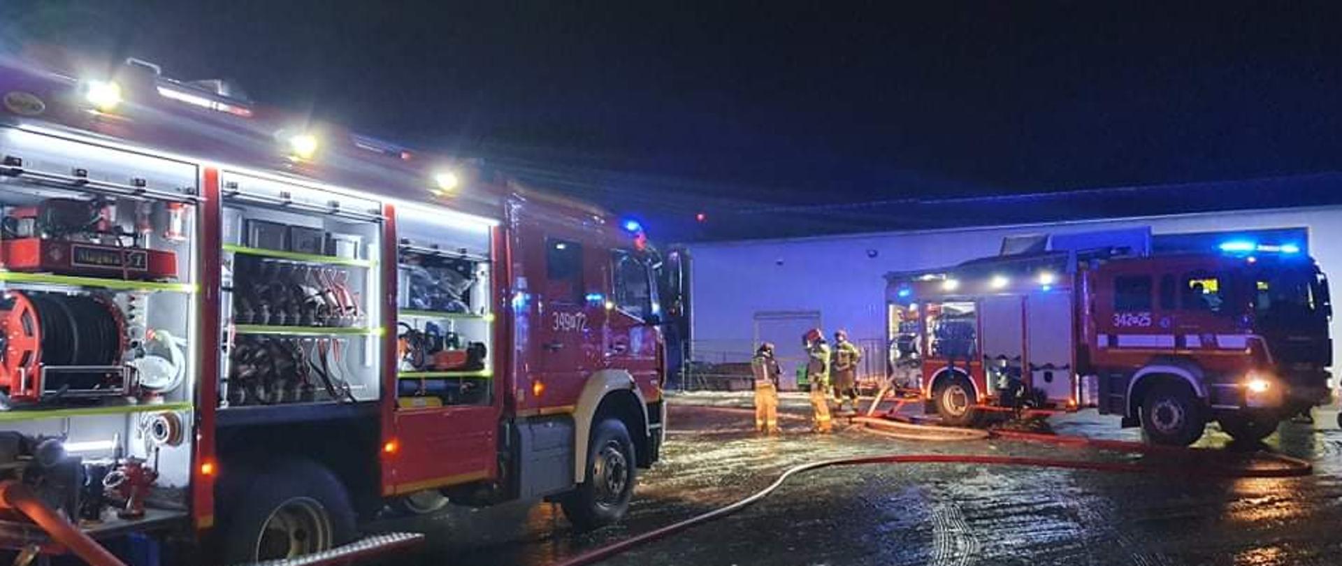 Na zdjęciu widać dwa pojazdy gaśnicze. Pojazdy posiadają kolor czerwone, białe oznakowanie i sygnalizację ostrzegawczą. Pora nocna. Pora roku to zima. W tle budynek w kolorze szarym. Pomiędzy autami strażak w ubraniu specjalnym w kolorze pisakowym. Posiada hełm w kolorze czerwonym. Auta mają włączone oświetlenie pola pracy.