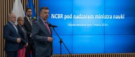 Minister Wieczorek i wiceministrowie stoją z boku sali, pod wielkim wbudowanym w ścianę ekranem z napisem NCBR pod nadzorem ministra nauki, minister mówi do mikrofonu na stojaku.