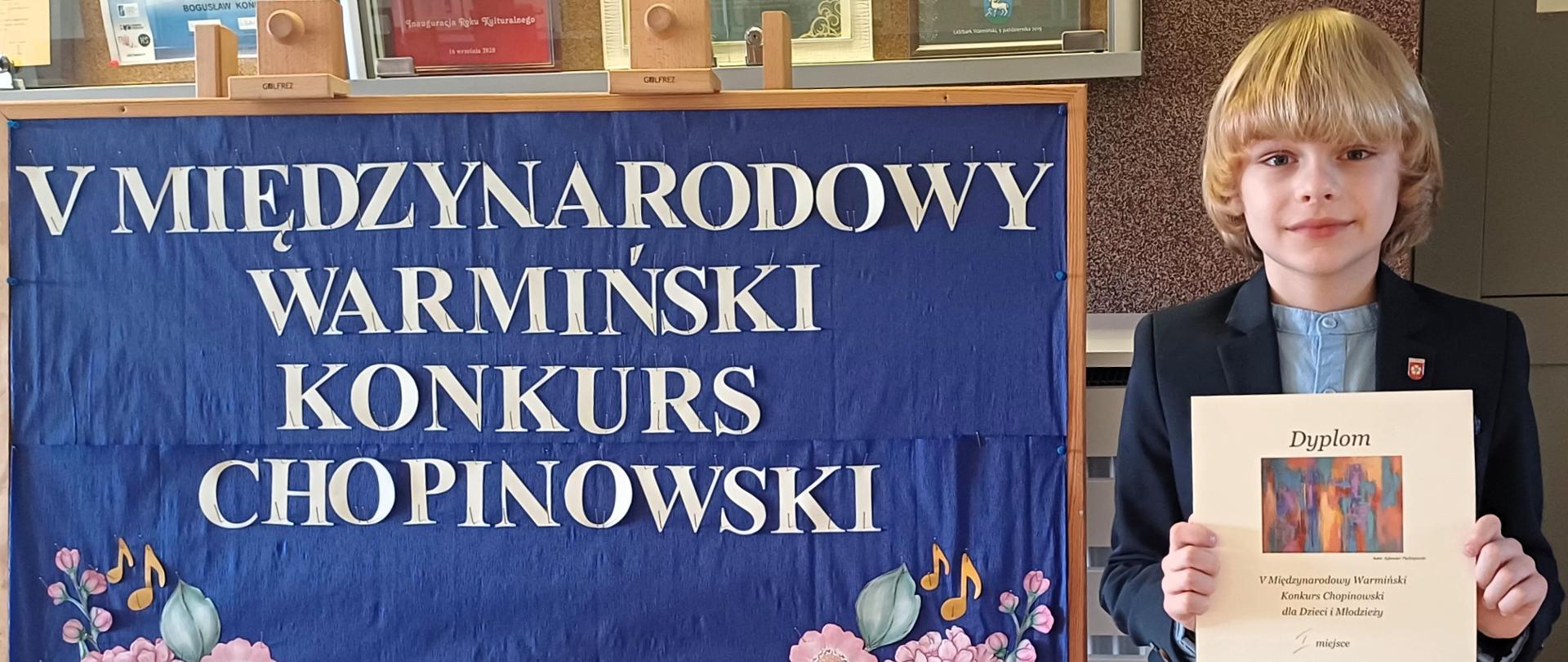 Po lewej niebieska tablica z nazwą konkursu, po prawej chłopiec z dyplomem