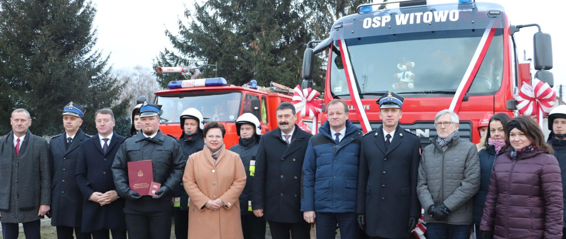 zdjęcie grupowe strazaków, kobiet i mężczyzn , w tle dwa samochody pożarnicze z biało czerwonymi wstęgami