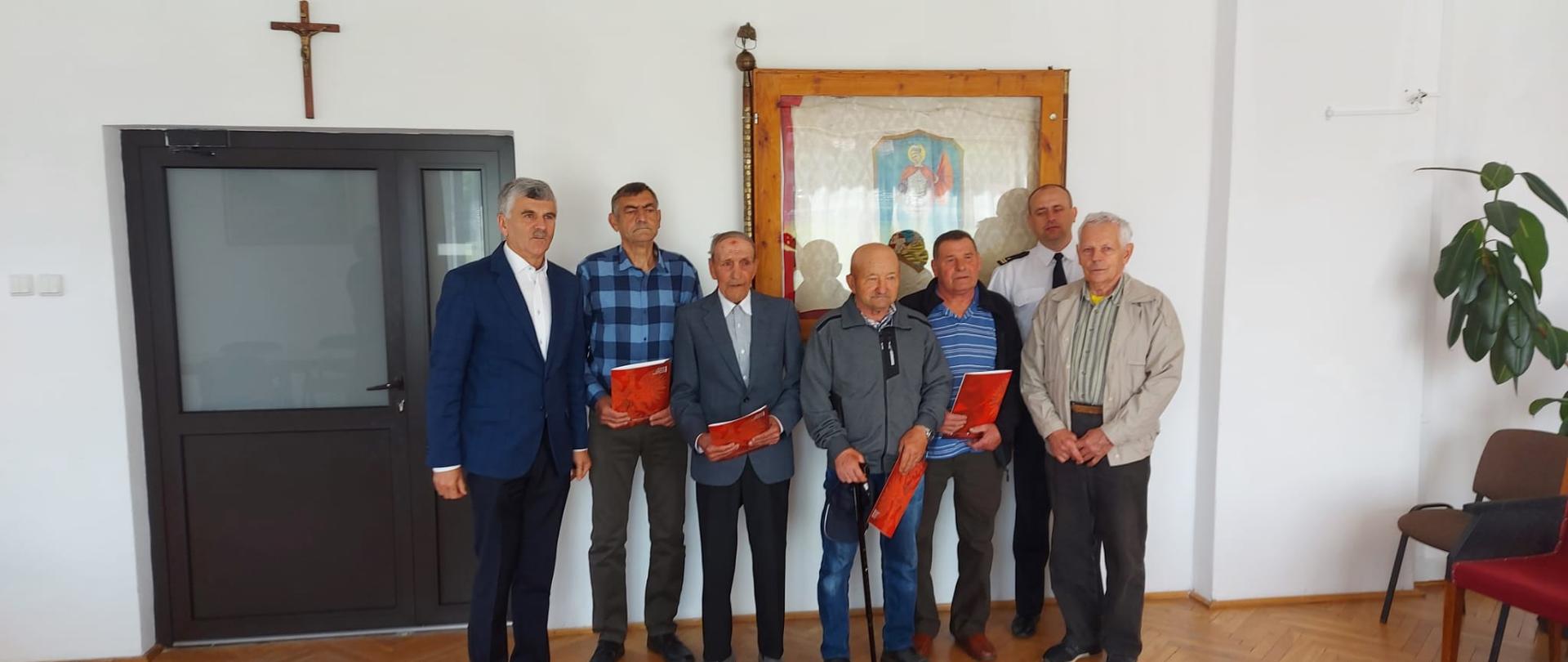 Na zdjęciu widać komendanta powiatowego PSP w Ustrzykach Dolnych wraz z wójtem gminy i pięcioma strażakami OSP którym wręczono świadczenia ratownicze. Komendant jest ubrany w mundur galowy. Za osobami znajduje się sztandar powieszony na ścianie i krzyż nad drzwiami. Strażacy trzymają w rękach teczki. 