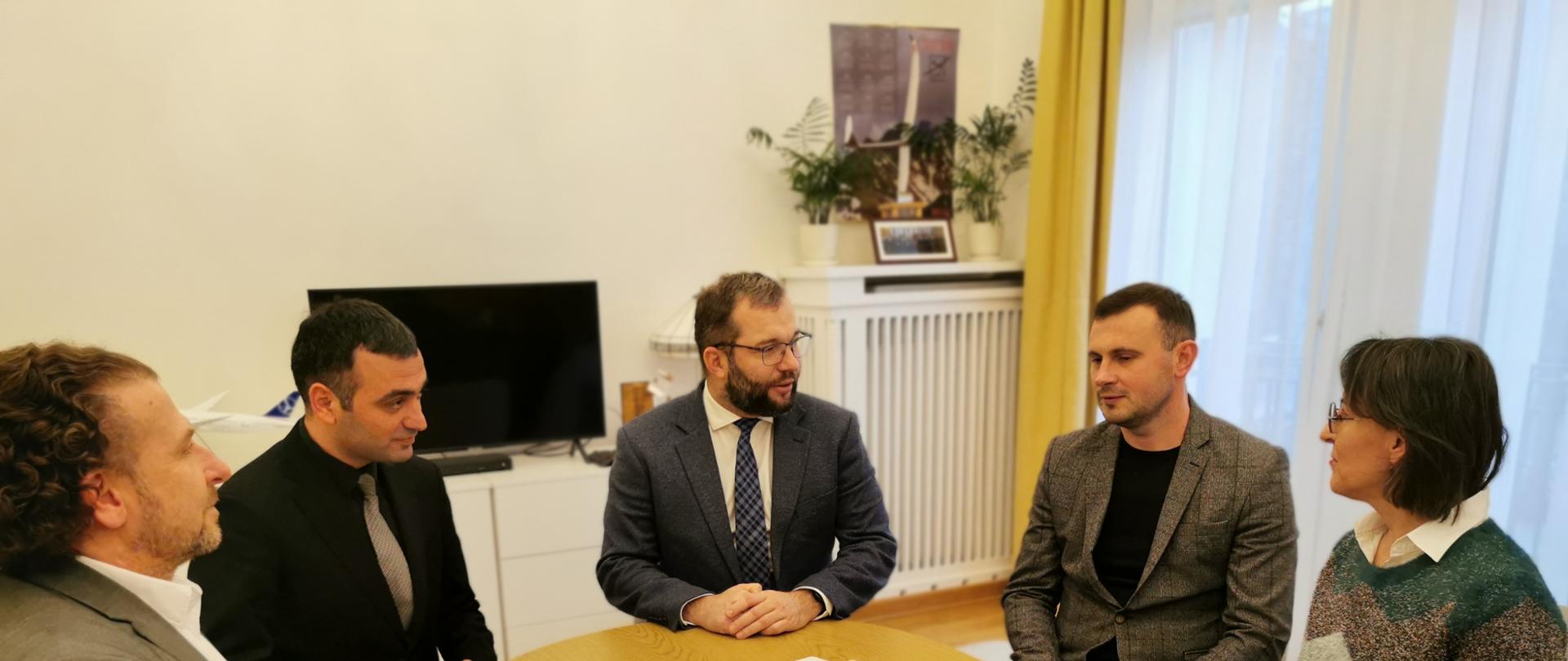 Czterech mężczyzn i kobieta siedzą przy prostokątnym stole z zaokrąglonymi rogami. Trzeci z lewej siedzi minister funduszy i polityki regionalnej.