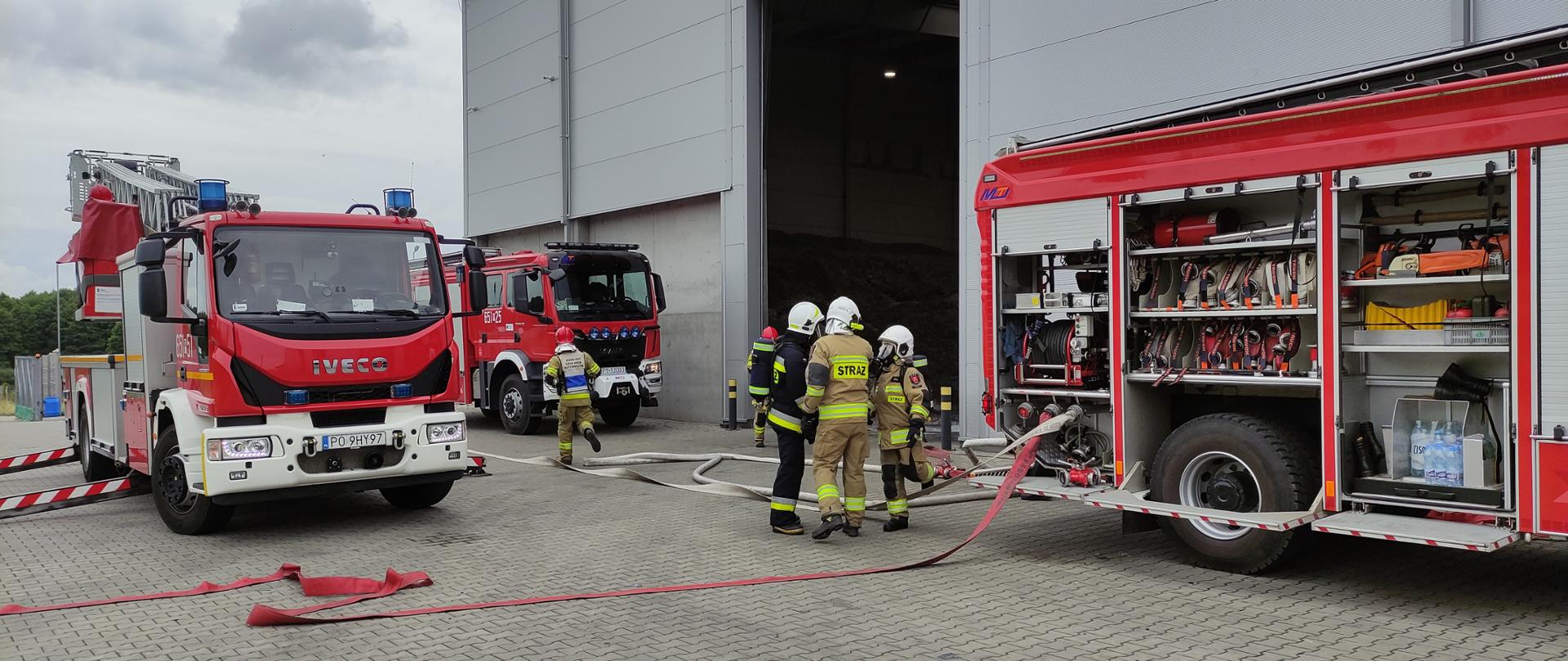 Zdjęcie przedstawia trzy wozy strażackie oraz strażaków stojących przed halą zakładu. Na ziemi rozwinięto węże pozarnicze