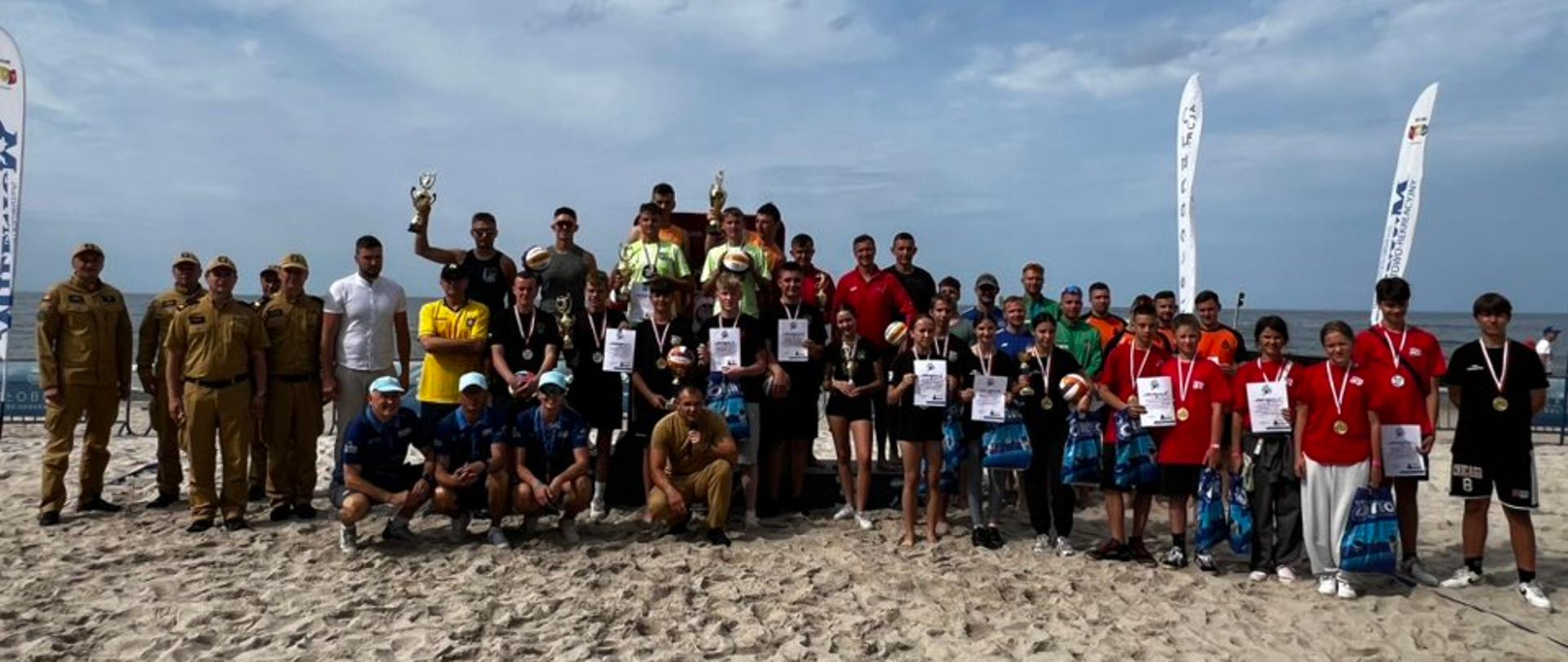 Na zdjęciu, zrobionym w słoneczny dzień, widać dużą grupę osób pozujących na piaszczystej plaży. W tle rozpościera się morze i błękitne niebo z białymi chmurami.
