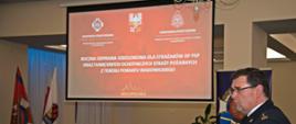 Widok z boku. Na środku ekran projekcyjny. Po lewej stronie ekranu znajdują się flagi Związku OSP RP, Państwowej Straży Pożarnej a w tle flaga biało – czerwona. Pod ekranem aranżacja ze sprzętu pożarniczego. Przemawia komendant powiatowy PSP w Wadowicach.