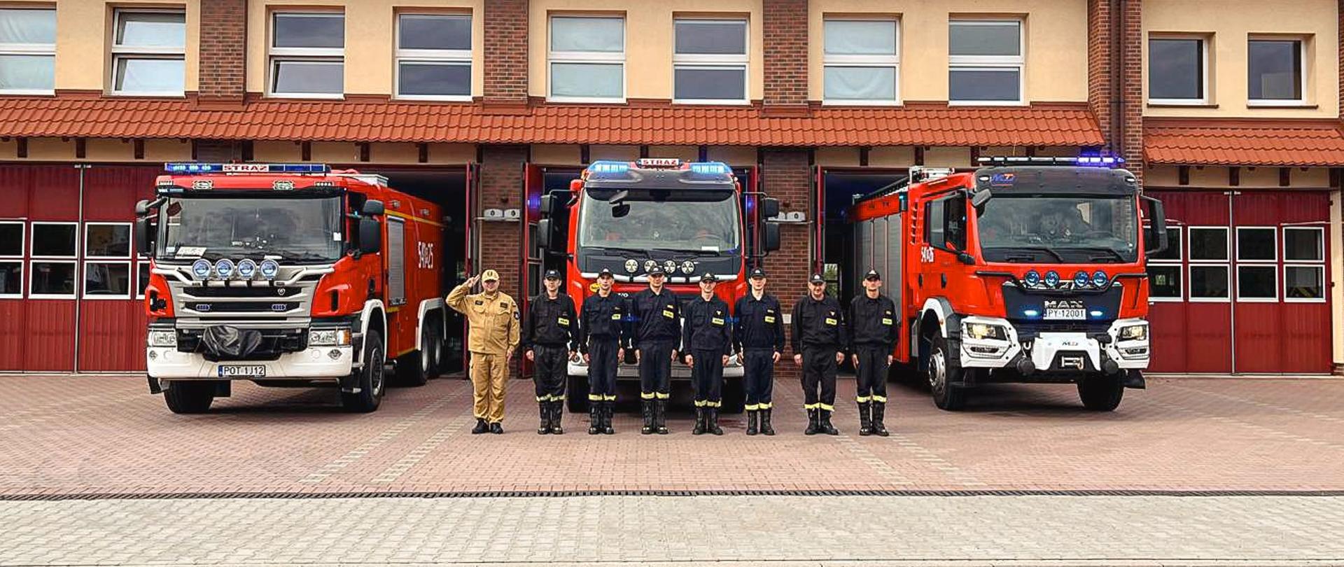 Strażacy stoją przed wozami strażackimi i strażnicą oddając honor.