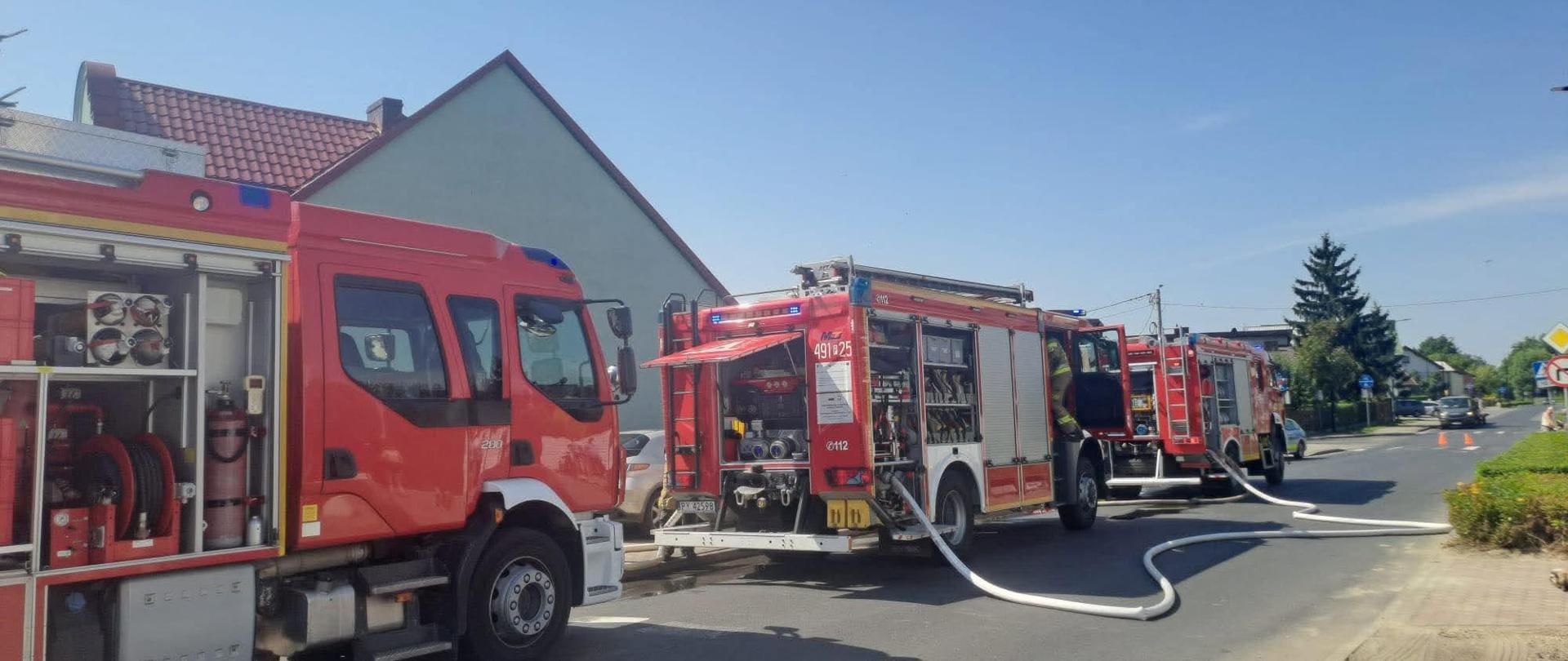 zdjęcie przedstawia pojazdy straży pożarnej podczas interwencji w związku z pozarem domu. Na ulicy widoczne również węże pożarnicze, którymi dostarczana jest woda do celów gaśniczych 