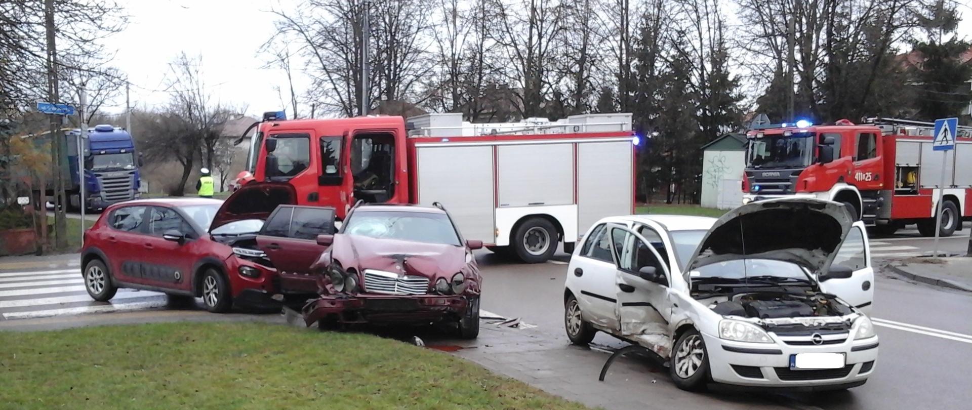 Rozbite trzy samochody osobowe stojące na chodniku przy skrzyżowaniu ulic Białowieskiej i Hołowieskiej w Bielsku Podlaskim. Pojazdy widoczne są od przodu. Od prawej strony: biały Opel Corsa, wiśniowy Mercedes E220, czerwony Citroen C3. W tle widoczne 2 samochody ratowniczo-gaśnicze z JRG w Bielsku Podlaskim. Na kolejnym planie widoczny jest funkcjonariusz Policji kierujący ruchem oraz samochód ciężarowy stojący w korku.