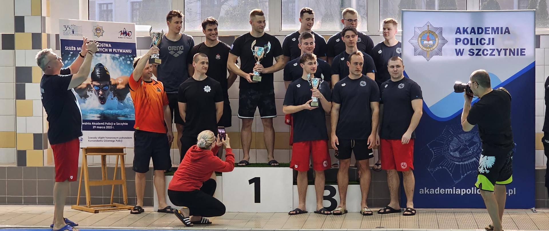 Zdjęcie przedstawia ceremonię wręczenia nagród na zawodach pływackich służb mundurowych, które odbyły się na pływalni Akademii Policji w Szczytnie. W centralnej części obrazu grupa młodych mężczyzn w sportowych koszulkach i spodenkach stoi na podium. Zawodnicy na najwyższym stopniu podium (oznaczonym numerem 1) trzymają w rękach puchary, symbolizujące ich zwycięstwo.
Po lewej stronie zdjęcia znajduje się mężczyzna w czarnej koszulce i czerwonych spodenkach, który podnosi ręce, prawdopodobnie ciesząc się z sukcesu drużyny. Przed nim, kobieta w czerwonej bluzie i czarnych spodniach kuca, robiąc zdjęcie telefonem. Po prawej stronie fotograf robi zdjęcia aparatem, stojąc boso przy krawędzi basenu.
W tle widoczny jest baner Akademii Policji w Szczytnie, a obok niego tablica informacyjna dotycząca zawodów. Na basenie widać tory oddzielone pływakami w kolorach niebieskim, żółtym i czerwonym.
