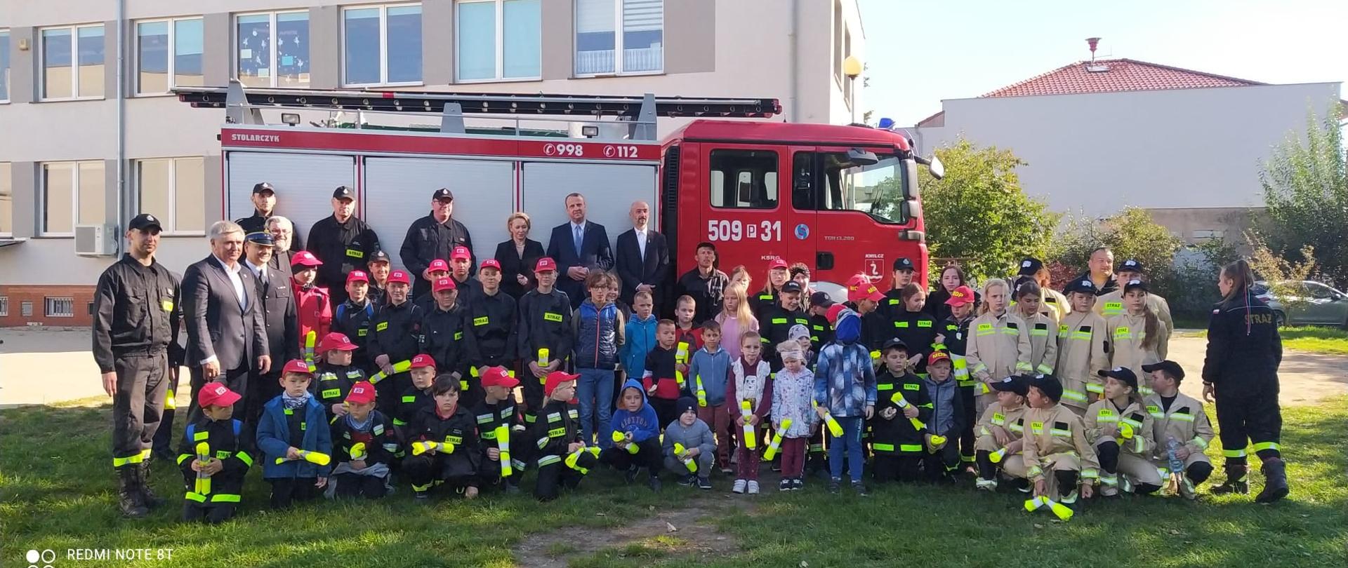 Zdjęcie grupowe uczestników rajdu i zaproszonych gości. W tle samochód pożarniczy.