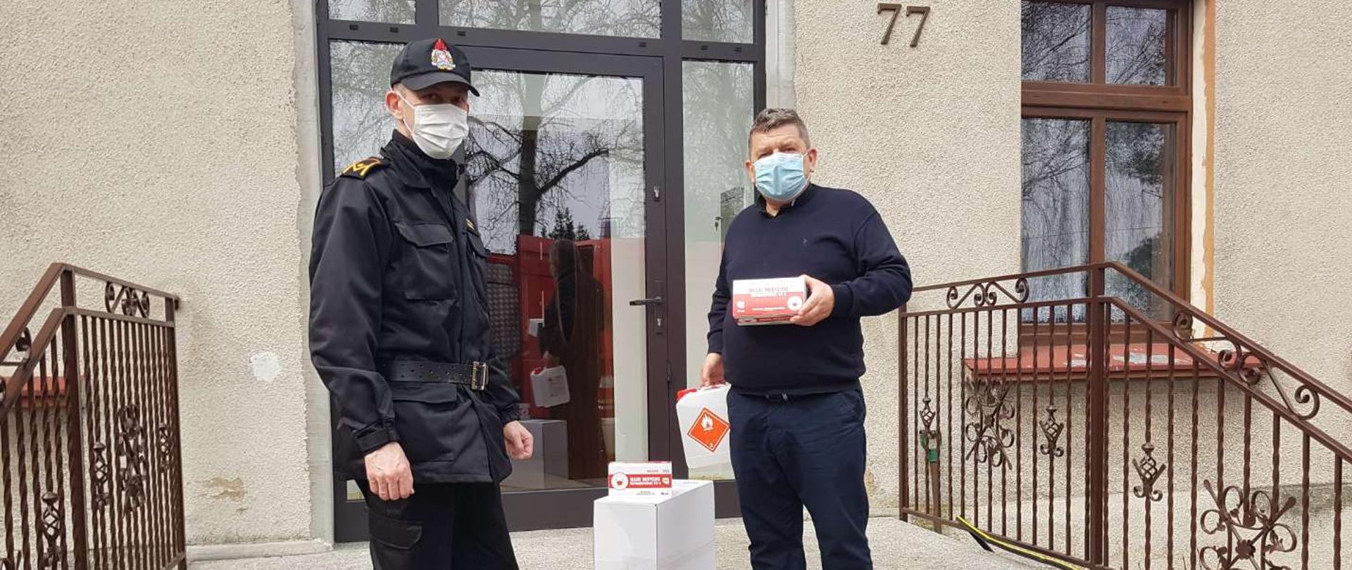Na zdjęciu widać po lewej stronie strażaka ubranego w czarny mundur typu KDS. Na twarzy biała maseczka. Na głowie czapka z daszkiem. Po prawej stronie osoba duchowna, ubrana w granatowy sweter. Na twarzy zielona maseczka. Osoba ta w ręku trzyma opakowanie z maskami ochronnymi koloru białego oraz w drugiej ręce pojemnik z płynem do dezynfekcji. Zdjęcie zrobione jest na schodach przed wejściem do kancelarii parafialnej. Za osobami przeszklone drzwi. Po bokach schodów metalowa barierka koloru brązowego.

