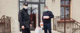 Na zdjęciu widać po lewej stronie strażaka ubranego w czarny mundur typu KDS. Na twarzy biała maseczka. Na głowie czapka z daszkiem. Po prawej stronie osoba duchowna, ubrana w granatowy sweter. Na twarzy zielona maseczka. Osoba ta w ręku trzyma opakowanie z maskami ochronnymi koloru białego oraz w drugiej ręce pojemnik z płynem do dezynfekcji. Zdjęcie zrobione jest na schodach przed wejściem do kancelarii parafialnej. Za osobami przeszklone drzwi. Po bokach schodów metalowa barierka koloru brązowego.
