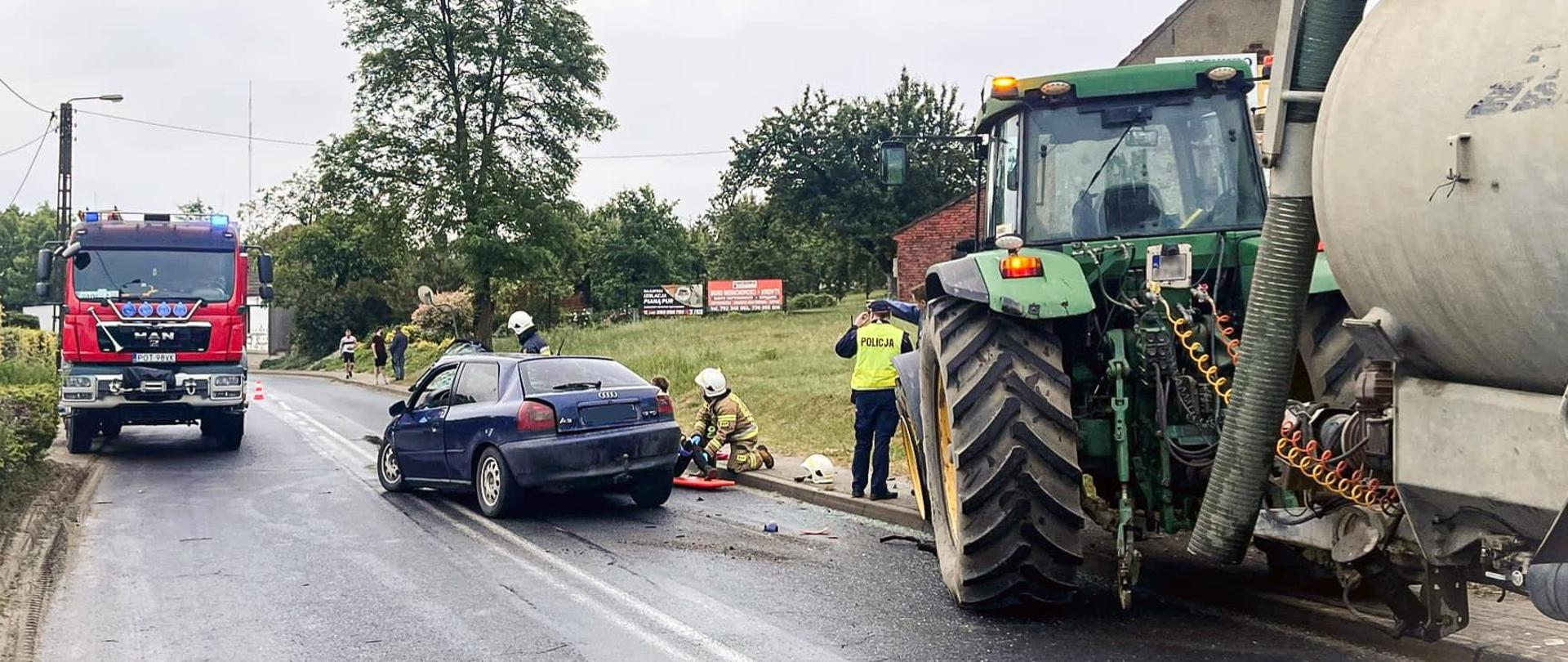Po prawej stronie traktor z beczką stojący na chodniku, na środku jezdni rozbity samochód osobowy, przy którym strażacy udzielają pomocy osobie poszkodowanej. Przed traktorem stoi policjant. Po lewej stronie wóz strażacki.