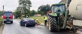 Po prawej stronie traktor z beczką stojący na chodniku, na środku jezdni rozbity samochód osobowy, przy którym strażacy udzielają pomocy osobie poszkodowanej. Przed traktorem stoi policjant. Po lewej stronie wóz strażacki.