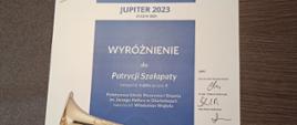 Dyplom na białym tle niebieski prostokąt. Napis Jupiter 2023 21 22 kwietnia 2023 poniżej białe litery wyróżnienie dla Patrycji Szałapaty kategoria trąbka grupa druga Państwowa Szkoła Muzyczna I Stopnia im Jerzego Hellera w Głuchołazach nauczyciel Władysław Wojtyła z prawej strony podpisy jurorów profesor doktor habilitowany Wacław Mulak doktor habilitowany Tomasz Stolarczyk magister Adam Kozłowski. Na dole widoczny fragment zdjęcia trąbki. 