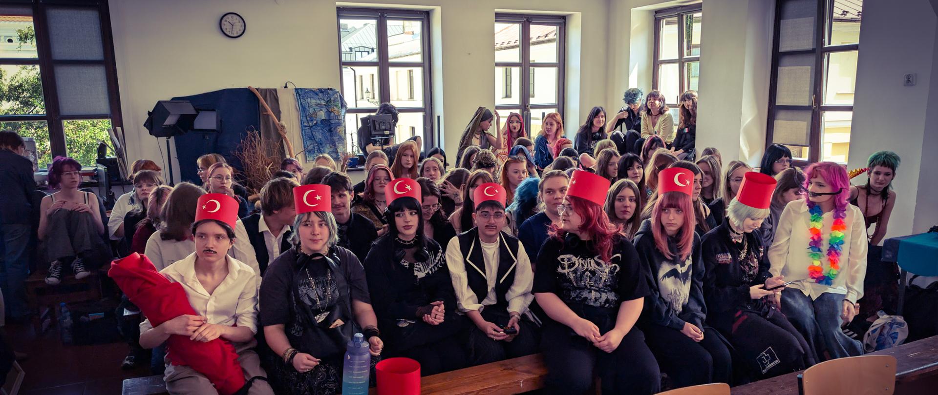 Zdjęcie przedstawia dużą grupę młodzieży zgromadzoną w sali lekcyjnej lub audytoryjnej. Wszyscy siedzą na drewnianych ławach i krzesłach, zwróceni w stronę obiektywu, co sugeruje widownię czekającą na wydarzenie lub uczestniczącą w spotkaniu.
Głównym punktem uwagi jest pierwszy rząd siedzących osób, z których większość ma na sobie czerwone, wysokie czapki (fezy) z białym półksiężycem i gwiazdą, nawiązujące do motywu tureckiego