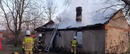 Na zdjęciu widać budynek ze spalonym dachem. Nad budynkiem szary dym. Zdjęcie zrobione w porze porannej. Wokół budynku drzewa bez liści. Przed budynkiem drabina, strażak w ubraniu specjalnym, wentylator osiowy.