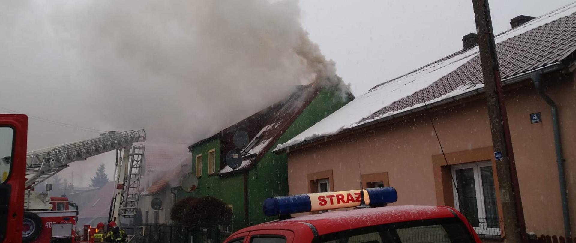 Zdjęcie przedstawia pożar dachu budynku mieszkalnego. Z dachu wydobywa się dym. Po lewej stronie zdjęcia widać samochód pożarniczy koloru czerwonego oraz pracujących na miejscu ratowników.