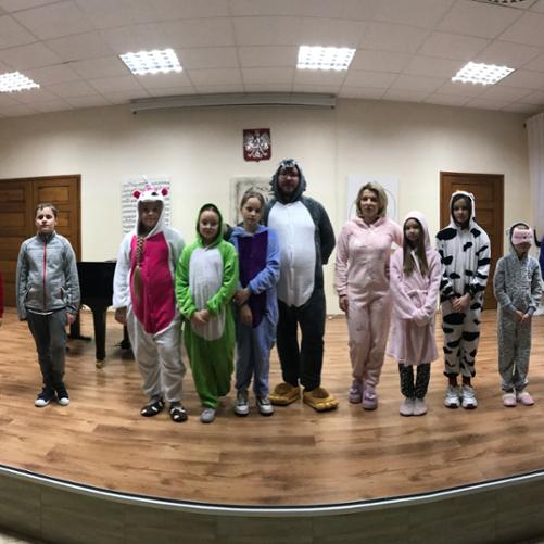 Uczniowie stoją na scenie w szeregu przebrani w piżamy, wraz z nimi nauczyciele Piotr Wilk w piżamie wilka oraz Ewelina Chrzanowska w piżamie jednorożca. 