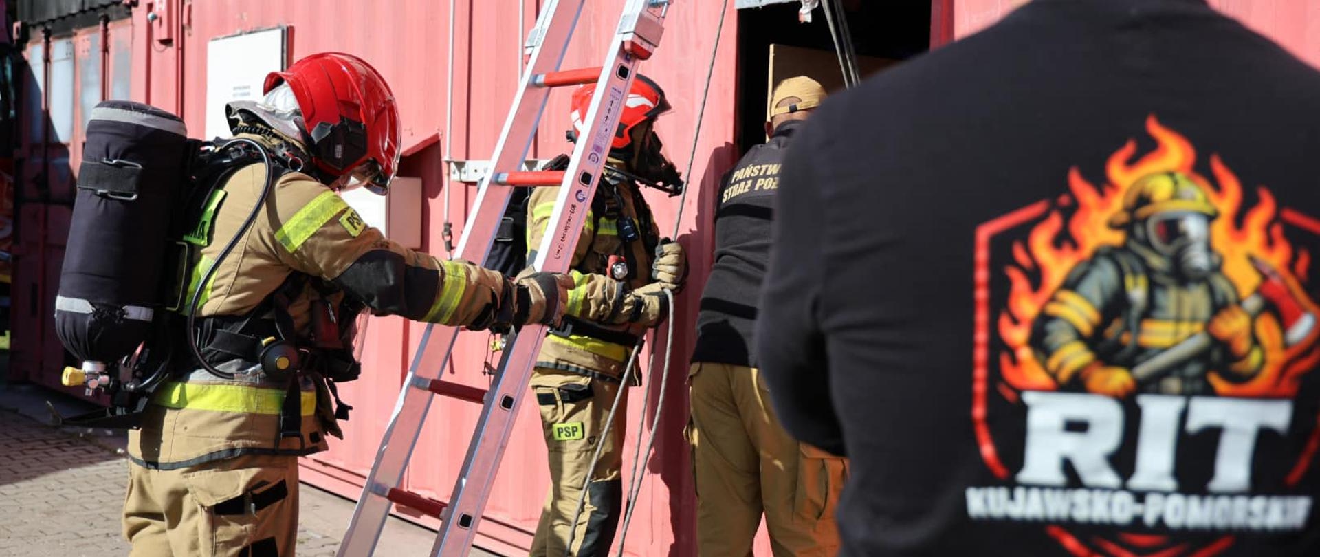 Strażak w ubraniu specjalnym i w aparacie ochrony dróg oddechowych stoi przy drabinie, opartej o hangar, za nimi inny strażak i strażak - sędzia. Z prawej strony stoi mężczyzna w koszulce symbolizującej Zawody RIT.