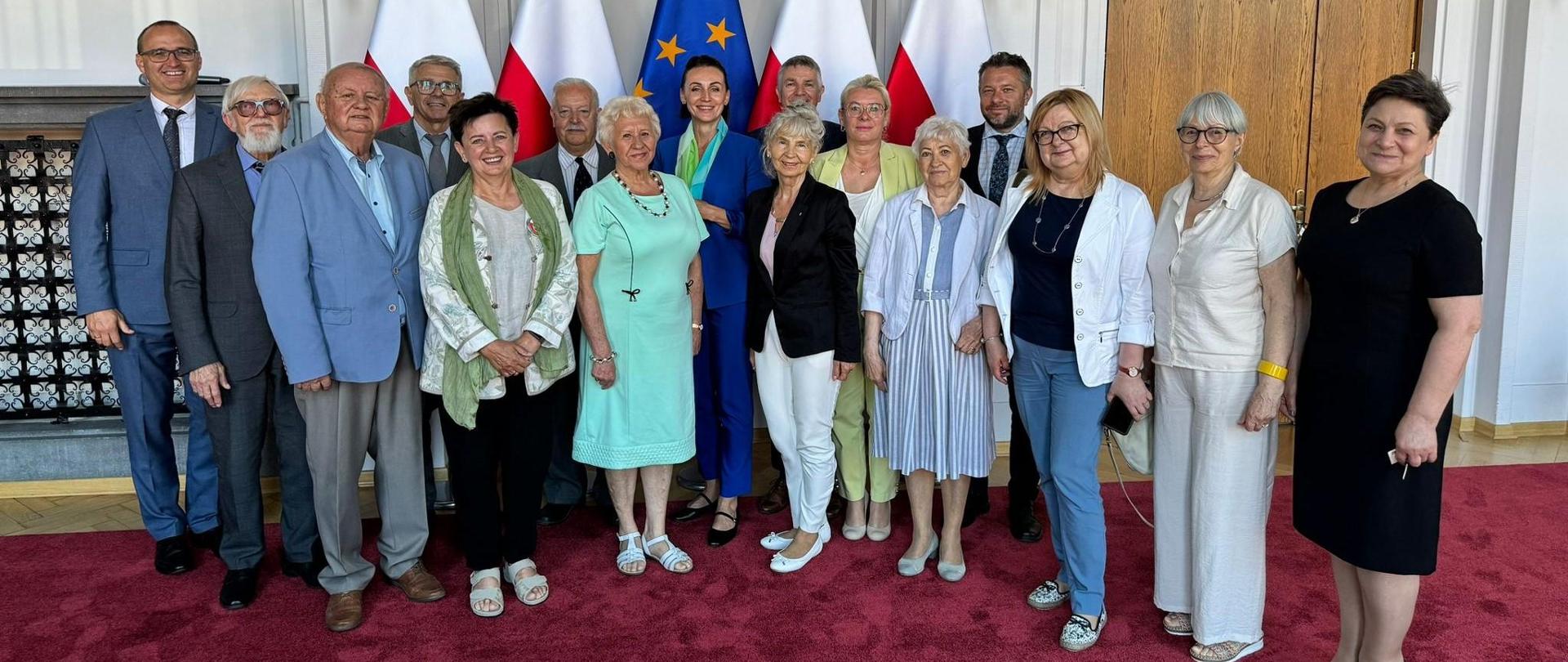 sala, grupa osób stoi na tle flag polskich i unijnych