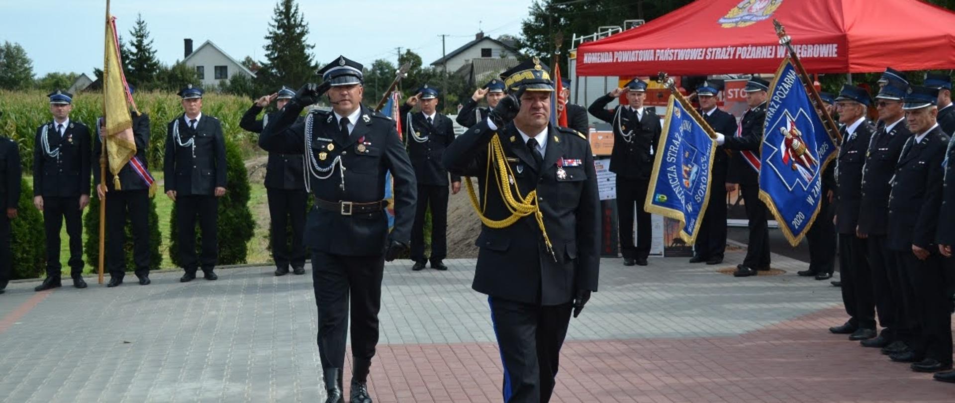 Mazowiecki Komendant Wojewódzki PSP nadbryg. Artur Gonera i przedstawiciel OSP oddają salut podczas uroczystego apelu jubileuszowego. W tle zgromadzeni strażacy, poczty sztandarowe oraz namiot Komendy Powiatowej PSP w Węgrowie.