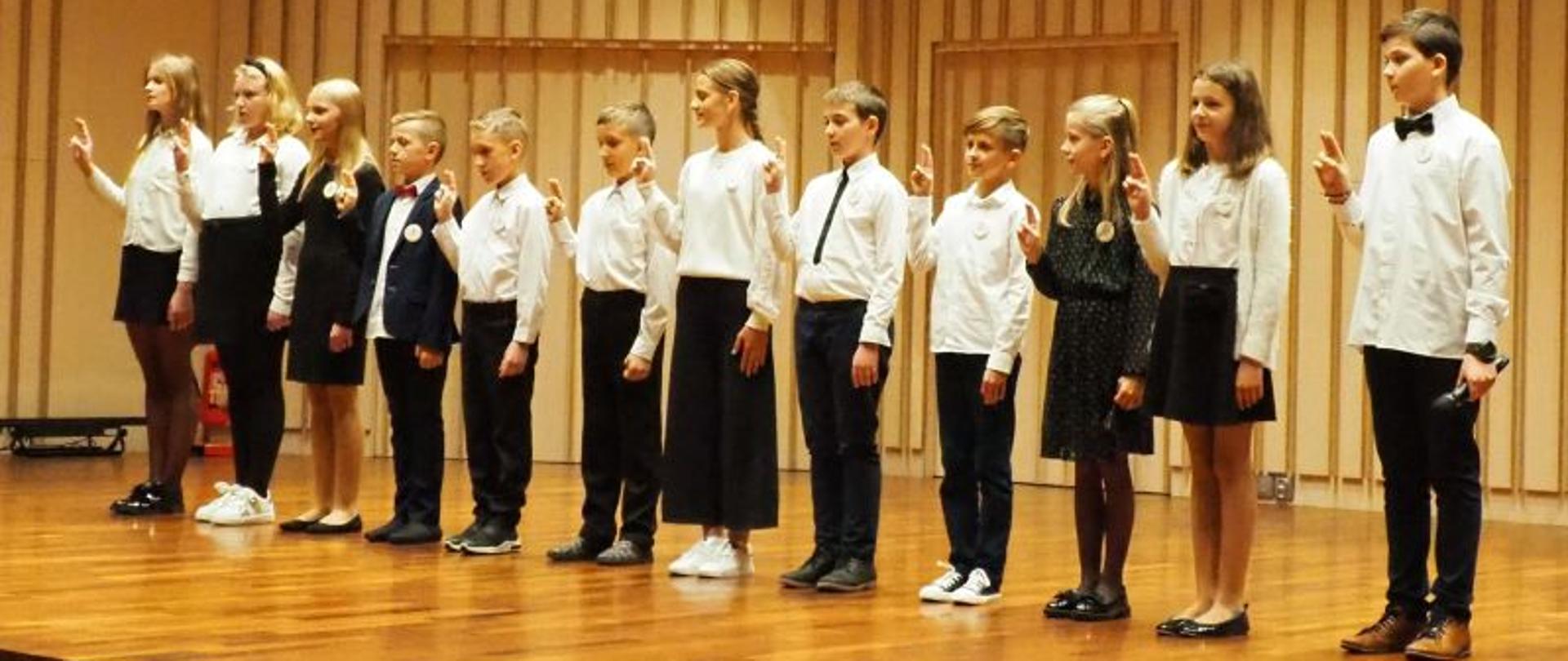 Dzieci klas pierwszych stoją na scenie w sali koncertowej PSM z podniesioną prawą dłonią, Dzieci ubrane na galowo