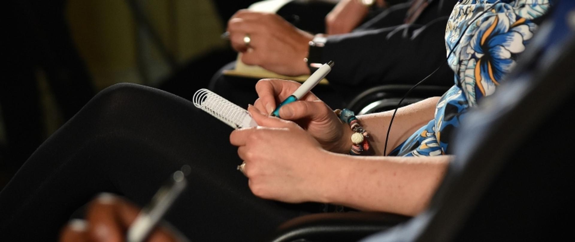 Osoby robiące notatki na notatnikach podczas spotkania lub szkolenia, widoczne dłonie i długopisy.
