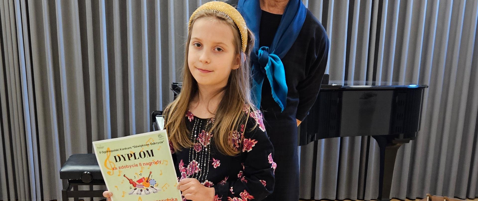 Uczennica z dyplomem i razem ze swoją Panią od instrumentu na tle fortepianu