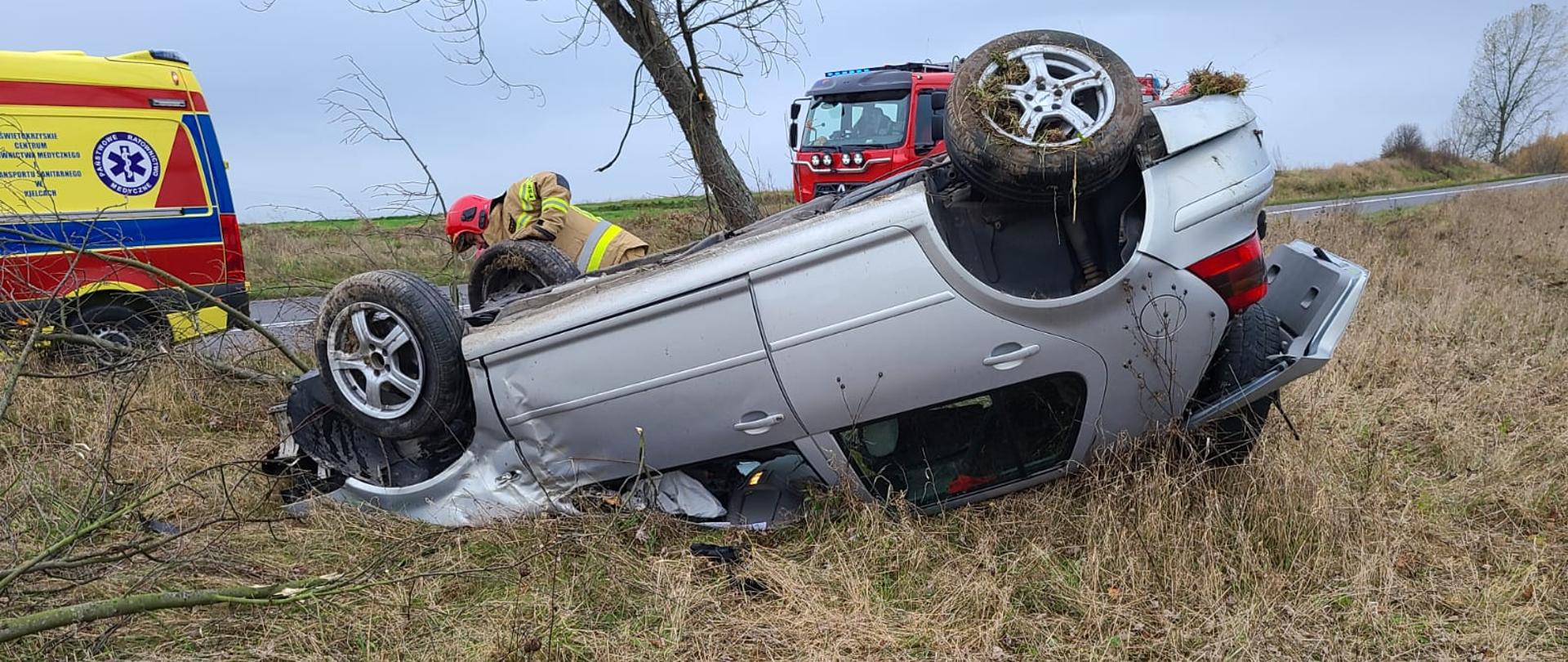 Zdjęcie przedstawia samochód osobowy leżący na poboczu na dachu. Za nim ratownik wykonuje czynności a na drodze powiatowej stoją ambulans i wóz strażacki.