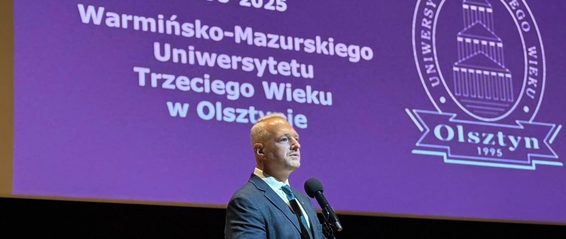 Mężczyzna stoi na scenie i przemawia, za nim wyświetlany jest obraz z napisami 