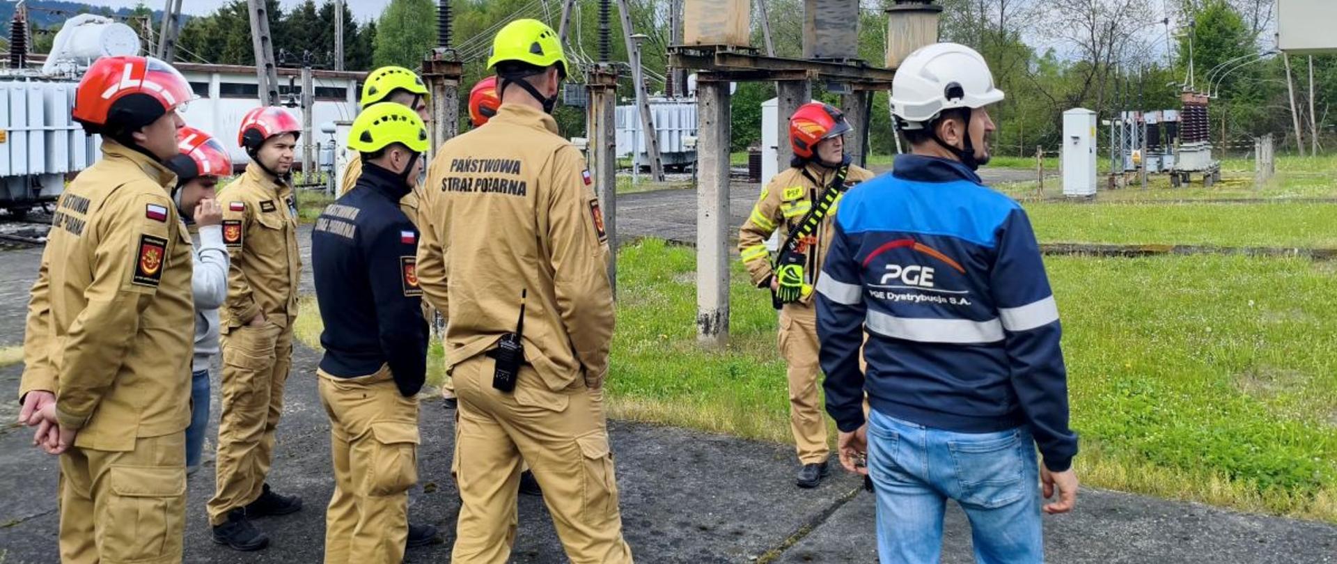 Na zdjęciu widać strażaków PSP w hełmach oraz pracownika PGE, którzy znajdują się na terenie stacji elektroenergetycznej PGE Dystrybucja S.A.
