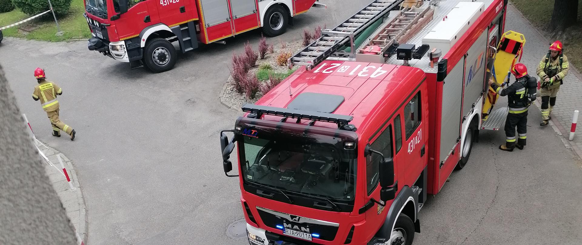 Na zdjęciu widzimy ustawione przed budynkiem szpitala dwa samochody pożarnicze, a także strażaków wyciągających z jednego z nich sprzęt ratowniczy. 