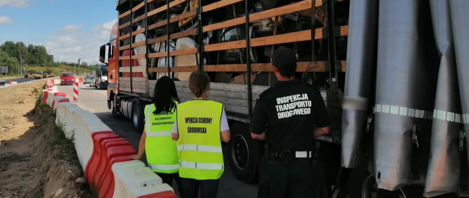 Na pierwszym planie: Inspektor ITD i pracownicy WIOŚ stoją obok naczepy z odsuniętą plandeką i kontrolują zawartość przestrzeni ładunkowej. W tle: oznakowany radiowóz ITD typu furgon i przebudowywana droga.