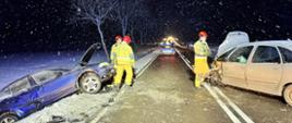 Nocne zdjęcie miejsca wypadku drogowego na zaśnieżonej trasie podczas opadów śniegu. Po lewej stronie, w przydrożnym rowie, znajduje się mocno uszkodzony niebieski samochód osobowy z podniesioną maską. Po prawej stronie, na pasie ruchu, stoi srebrny samochód z całkowicie rozbitym przodem. Na środku drogi pracują trzej strażacy w żółto-piaskowych mundurach i czerwonych hełmach. W tle widoczne są światła radiowozu oraz inne pojazdy służb ratunkowych. Jezdnia jest mokra, a pobocza pokryte grubą warstwą śniegu.