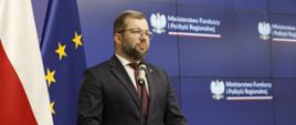 Minister funduszy i polityki regionalnej Grzegorz Puda stoi przy mikrofonie i przemawia, za jego plecami baner MFiPR 
