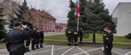 Trzech strażaków stoi przy maszcie, jeden z nich podnosi flagę. Po obu stronach masztu, naprzeciwko siebie stoją strażacy z dwóch zmian służbowych. Dowódca jednostki przeprowadza uroczystą zmianę służby z podniesieniem flagi państwowej.
