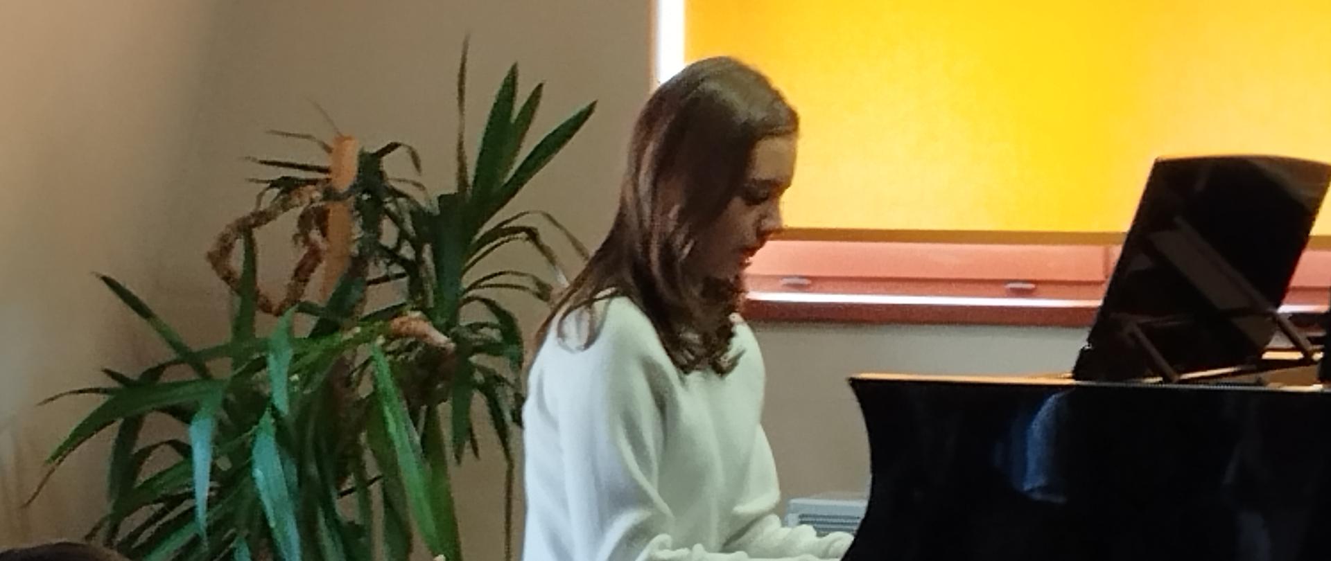 Na zdjęciu znajduje się młoda dziewczyna grająca na fortepianie podczas występu w sali muzycznej. Dziewczyna siedzi przy dużym, czarnym fortepianie koncertowym. Ubrana jest w prosty, biały sweter i ciemne spodnie. Ma długie, rozpuszczone włosy i pochylona jest lekko nad klawiaturą fortepianu, skupiona na grze. Jej postawa i wyraz twarzy sugerują pełne zaangażowanie i koncentrację na wykonywanym utworze.W tle znajduje się duża doniczkowa roślina o długich, wąskich liściach. Za nią widoczne jest okno przysłonięte pomarańczową roletą, przez którą wpada delikatne, ciepłe światło, nadając pomieszczeniu przytulny klimat. Na pierwszym planie, podobnie jak na poprzednim zdjęciu, widać tyły głów kilku słuchaczy, co wskazuje, że dziewczyna gra przed publicznością. Cała scena emanuje spokojem, skupieniem i muzyczną pasją.