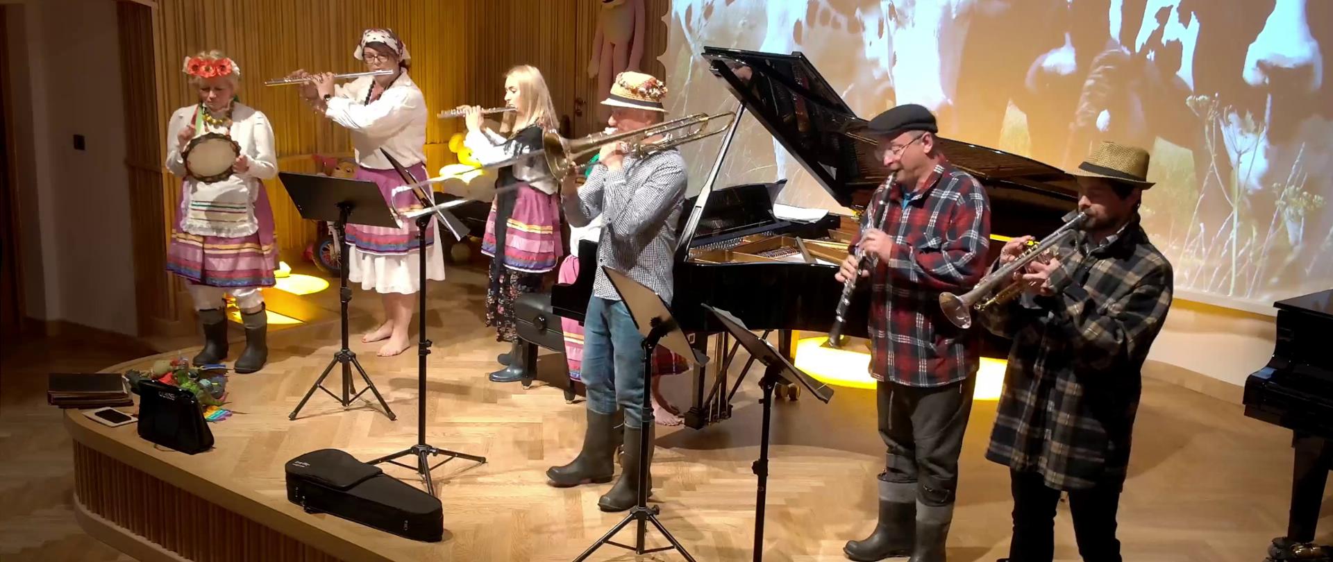 Zdjęcie. Na estradzie sali koncertowej występuje zespół instrumentów dętych. Wykonawcy to nauczyciele szkoły przebrani za mieszkańców wsi. od lewej wykonawcy: perkusistka (gra na tamburynie), dwie flecistki, puzonista, klarnecista i trębacz. Za nimi stoi otwarty czarny fortepian. W tle wyświetlony jest obraz przedstawiający stado krów.