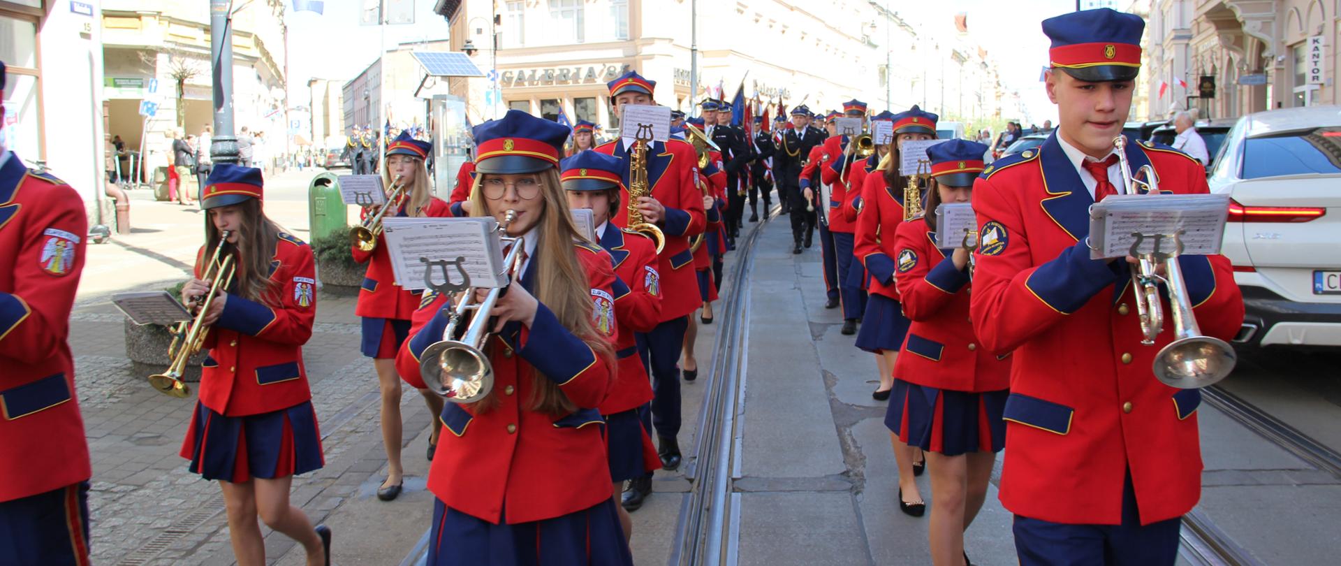 Widok na orkiestrę dętą. Orkiestranci w czerwono-granatowych mundurach idą ulicami miasta.