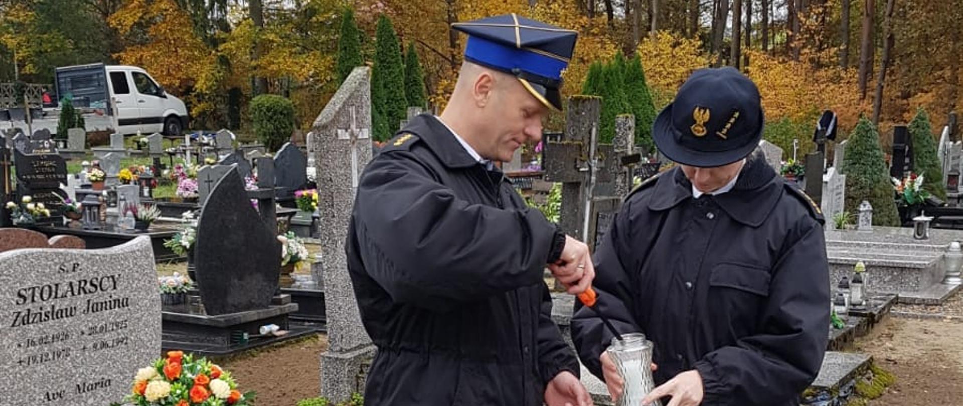 Zdjęcie przedstawia mężczyznę i kobietę w mundurach strażackich zapalających znicz. Stója przed grobem, oddając hołd zmarłym.